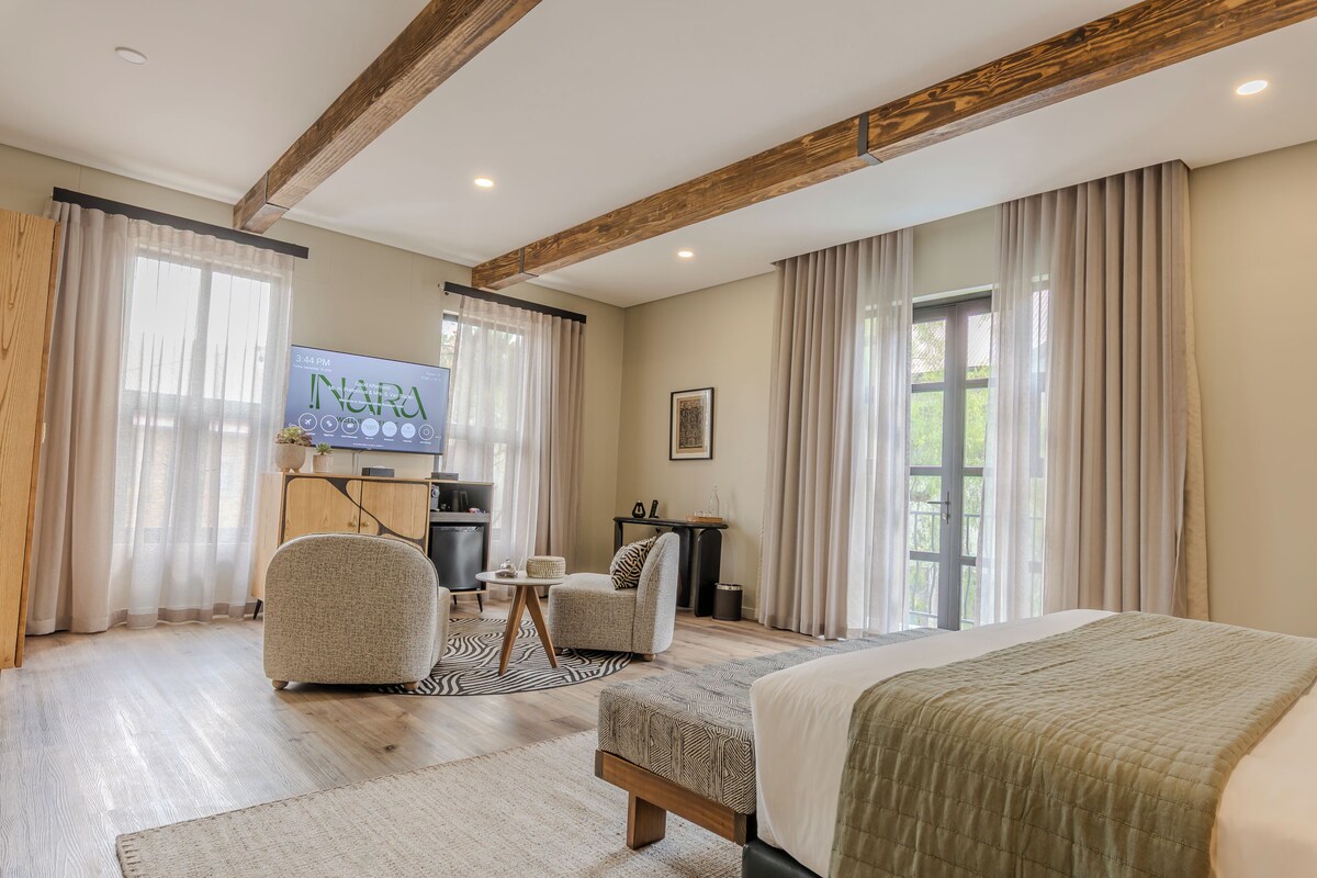 Une chambre d’hôtel comprenant un lit, deux fauteuils, une petite table, un téléviseur et une grande fenêtre avec des rideaux.