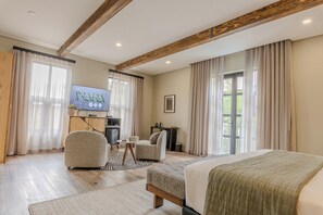 Une chambre d’hôtel comprenant un lit, deux fauteuils, une petite table, un téléviseur et une grande fenêtre avec des rideaux.