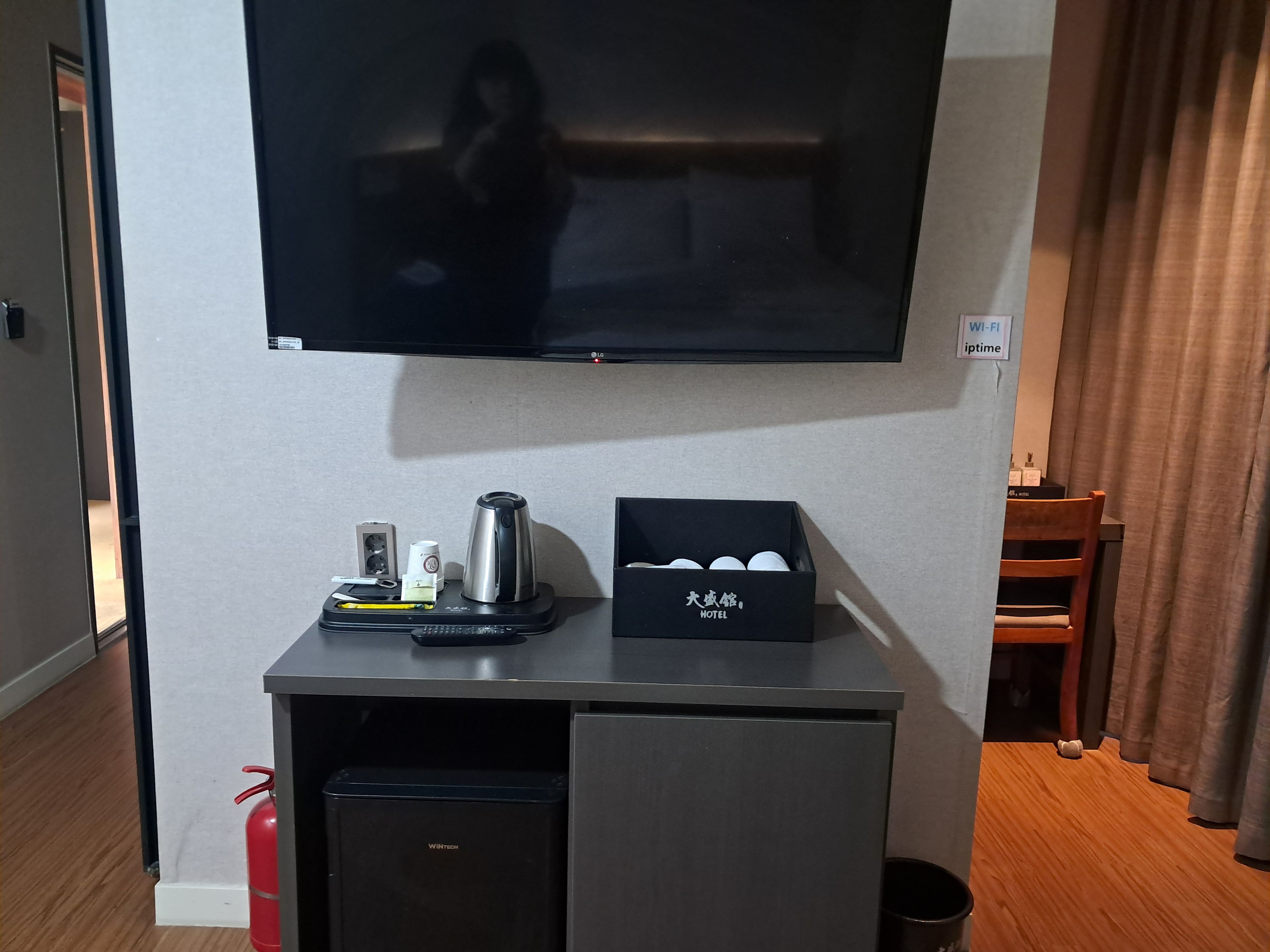 A hotel room with a flat-screen TV mounted on the wall, a small table with a kettle, a box of toiletries, and a compact refrigerator.
