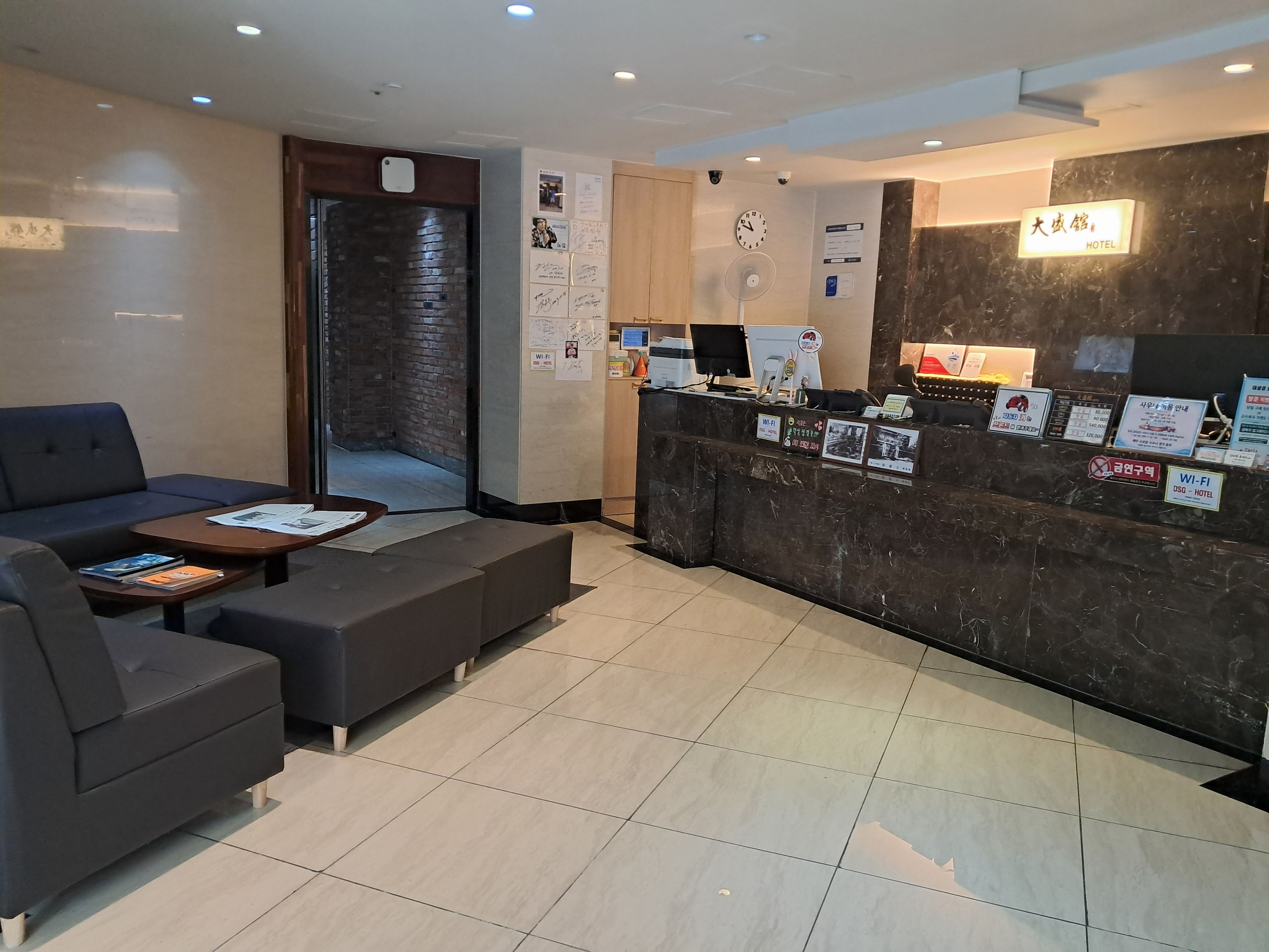 A hotel reception area with a dark reception desk, a seating area with sofas and a coffee table, and a clock on the wall.