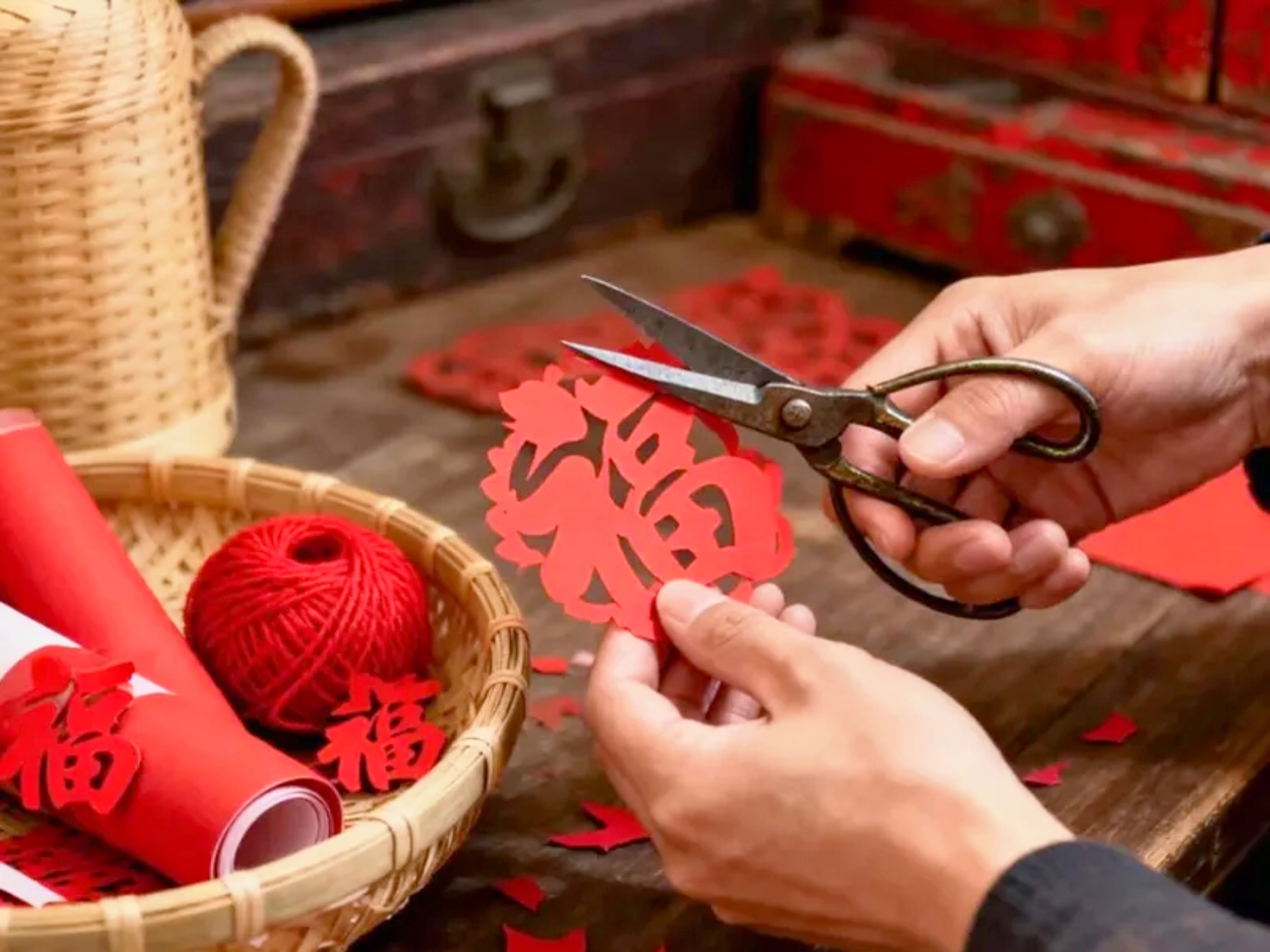 Manos cortando papel rojo con tijeras, un ovillo de lana roja y una cesta con rollos de papel rojo sobre una superficie de madera.