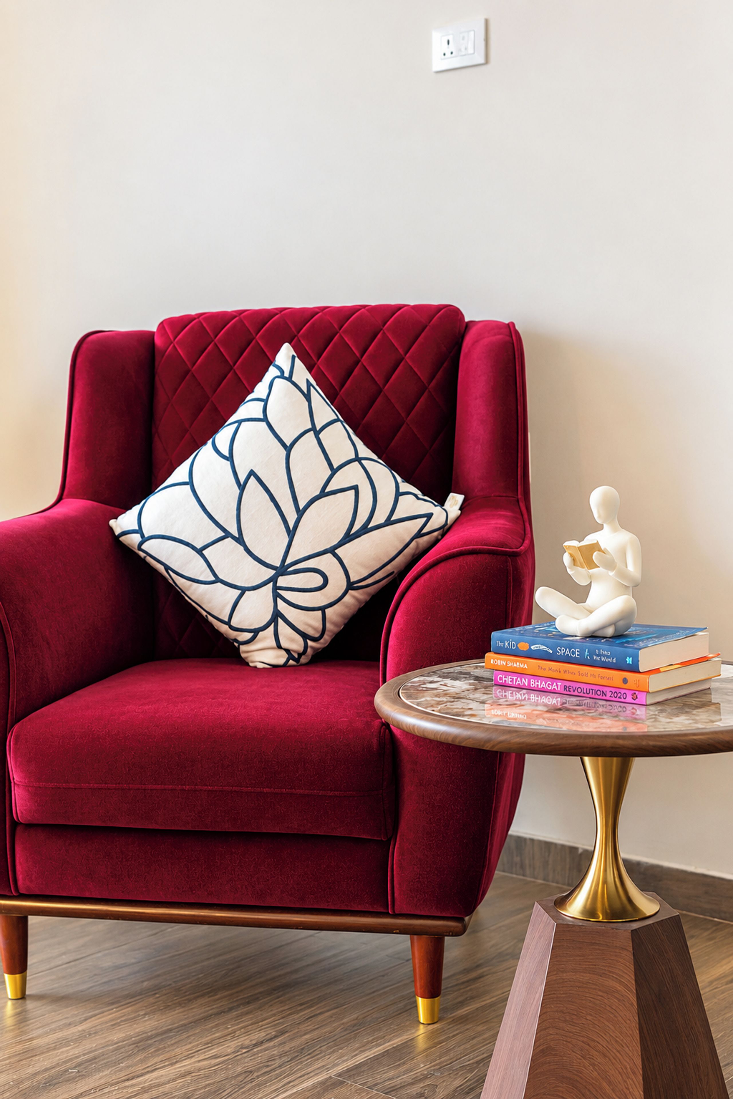 A red velvet armchair with a white and blue patterned pillow, a small white statue, and a stack of books on a round table.