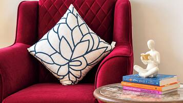 A red velvet armchair with a white and blue patterned pillow, a small white statue, and a stack of books on a round table.