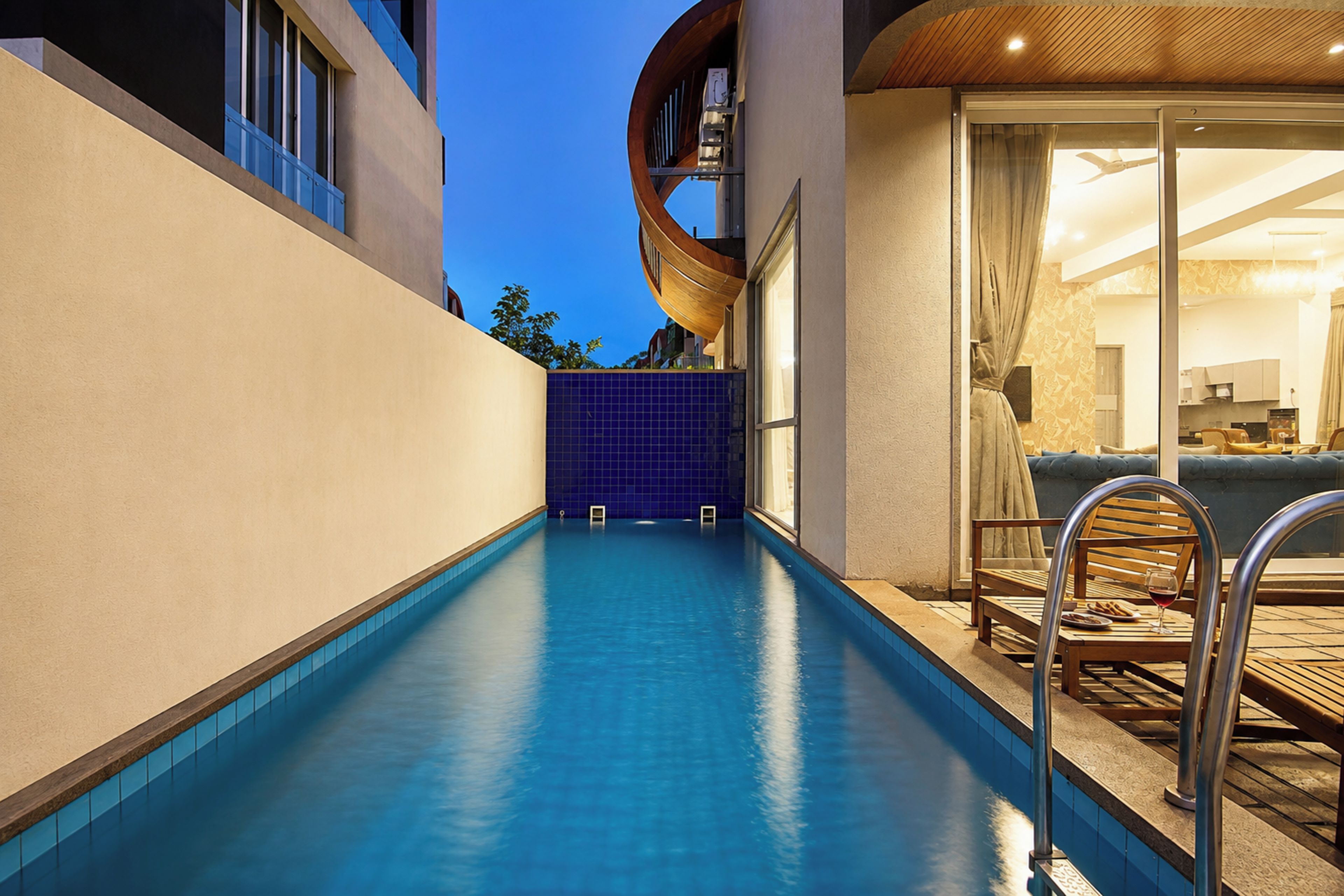 A modern indoor pool with clear blue water, a wooden deck, and a glass door leading to a living area with a sofa and a dining table.