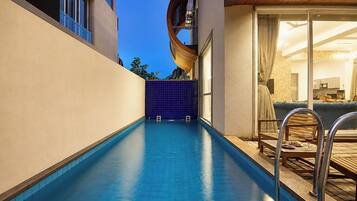 A modern indoor pool with clear blue water, a wooden deck, and a glass door leading to a living area with a sofa and a dining table.