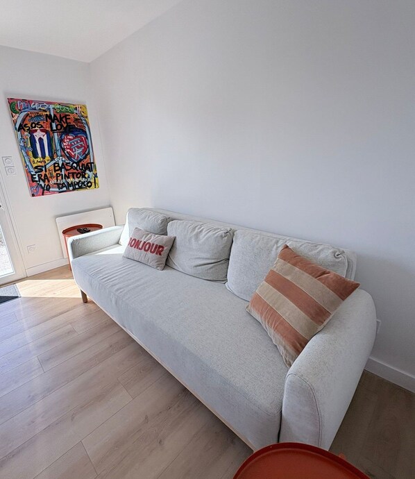 A light-colored sofa with two pillows, one with 'BONJOUR' text, in a room with a wooden floor and a colorful graffiti artwork on the wall.