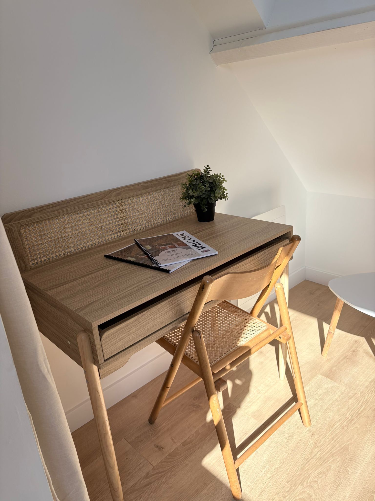 A wooden desk with a woven chair, a small potted plant, and a magazine on the table.
