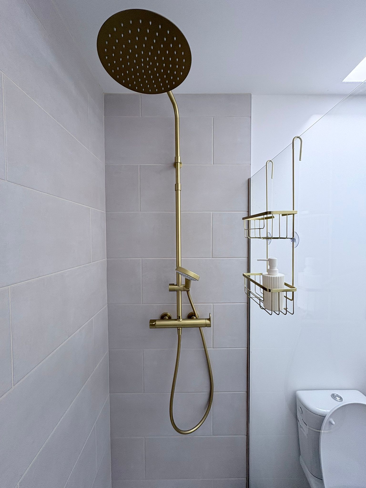 A shower with a gold showerhead and hose, a wall-mounted shelf with toiletries, and a toilet in a bathroom with light grey tiles.