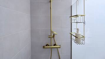 A shower with a gold showerhead and hose, a wall-mounted shelf with toiletries, and a toilet in a bathroom with light grey tiles.