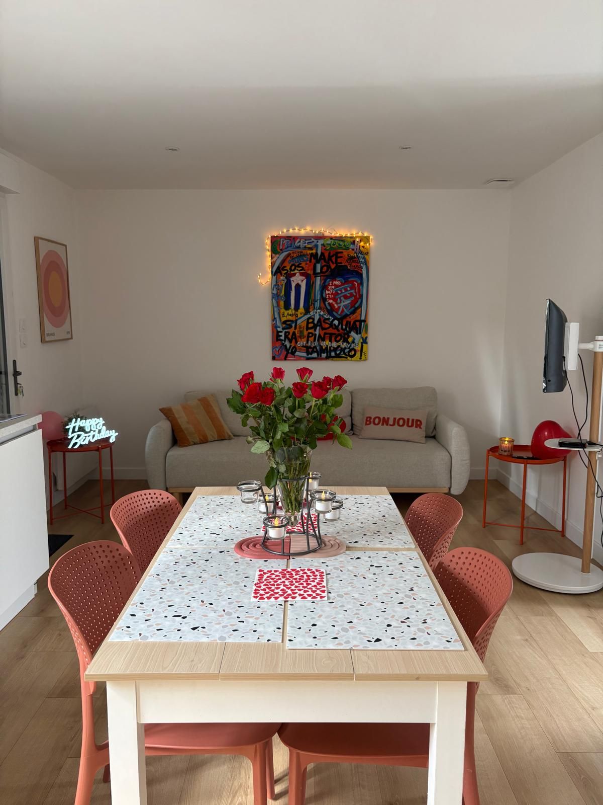 A dining area with a table set for four, surrounded by red chairs, a vase with red roses, and a wall-mounted artwork.