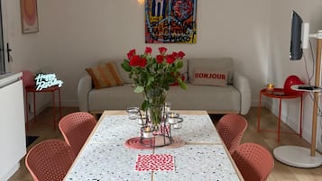 A dining area with a table set for four, surrounded by red chairs, a vase with red roses, and a wall-mounted artwork.