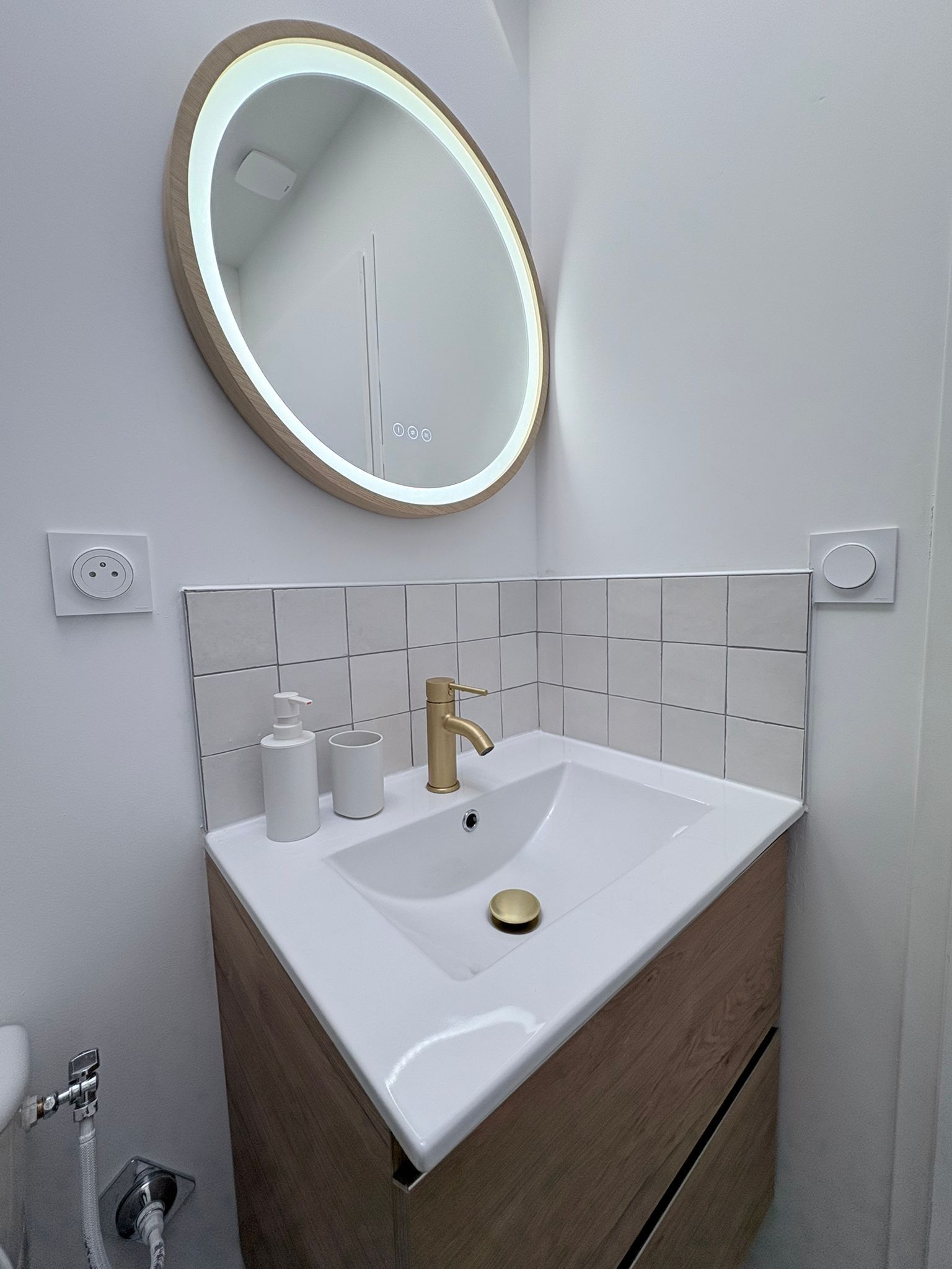 A bathroom with a white sink, gold faucet, and a round mirror with a light.