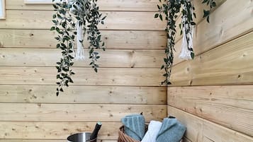 A wooden cabin interior with a wooden bench, a metal bucket, a bottle, a basket with towels, and hanging plants.