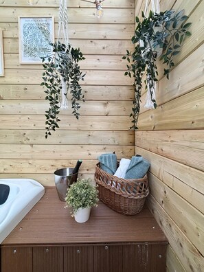 Interior d'una cabana de fusta amb una banca de fusta, un cubell metàl·lic, una ampolla, una cistella amb tovalloles i plantes penjants.