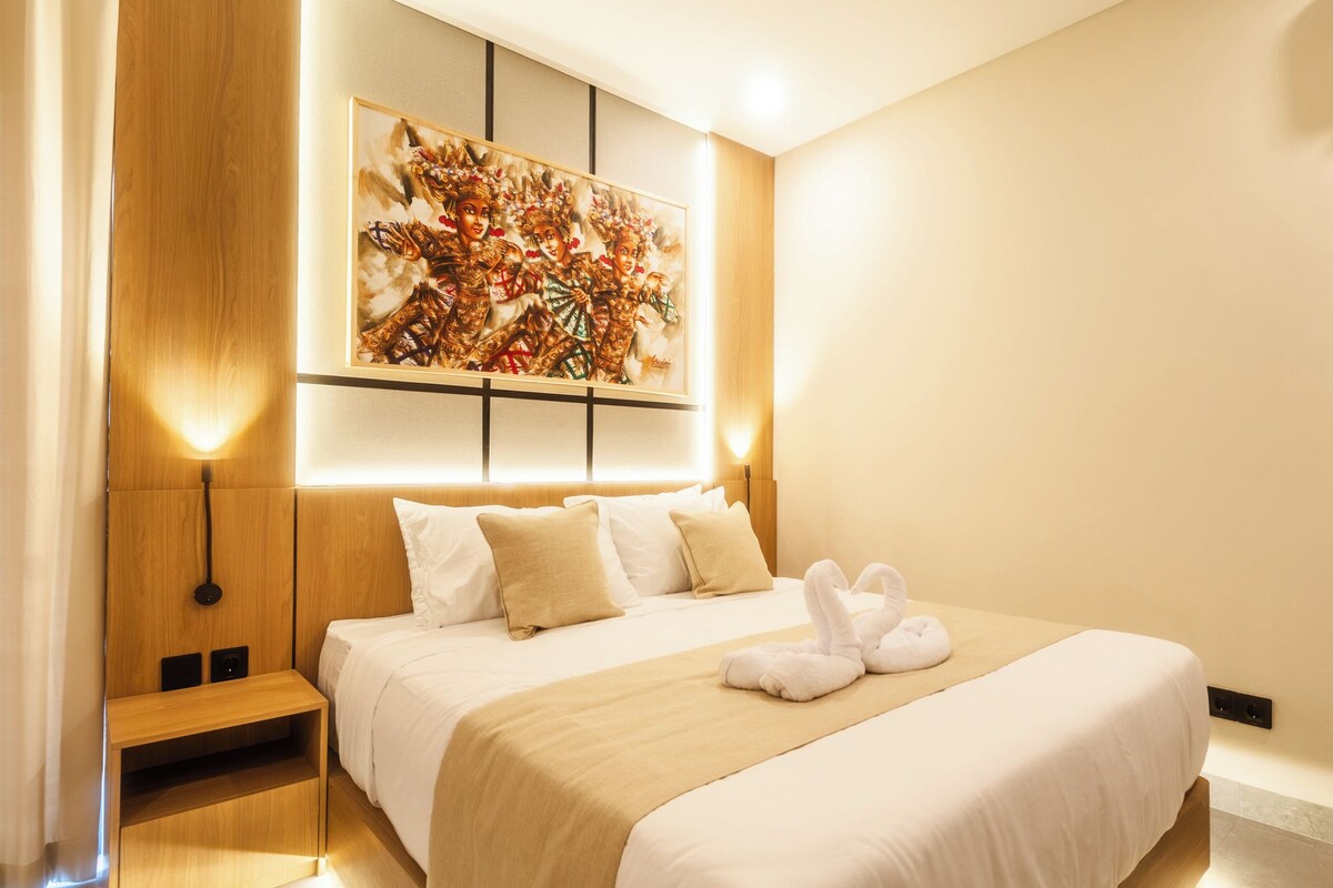 Una habitación de hotel moderna con una cama grande, un cabecero con iluminación integrada, una obra de arte colgada en la pared y una mesita de noche con lámpara.