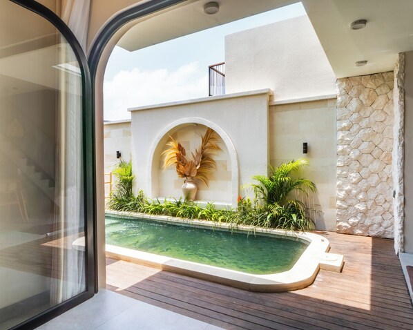 Un área de piscina con muro de piedra, terraza de madera y vista a un edificio con fachada blanca y ventana arqueada.