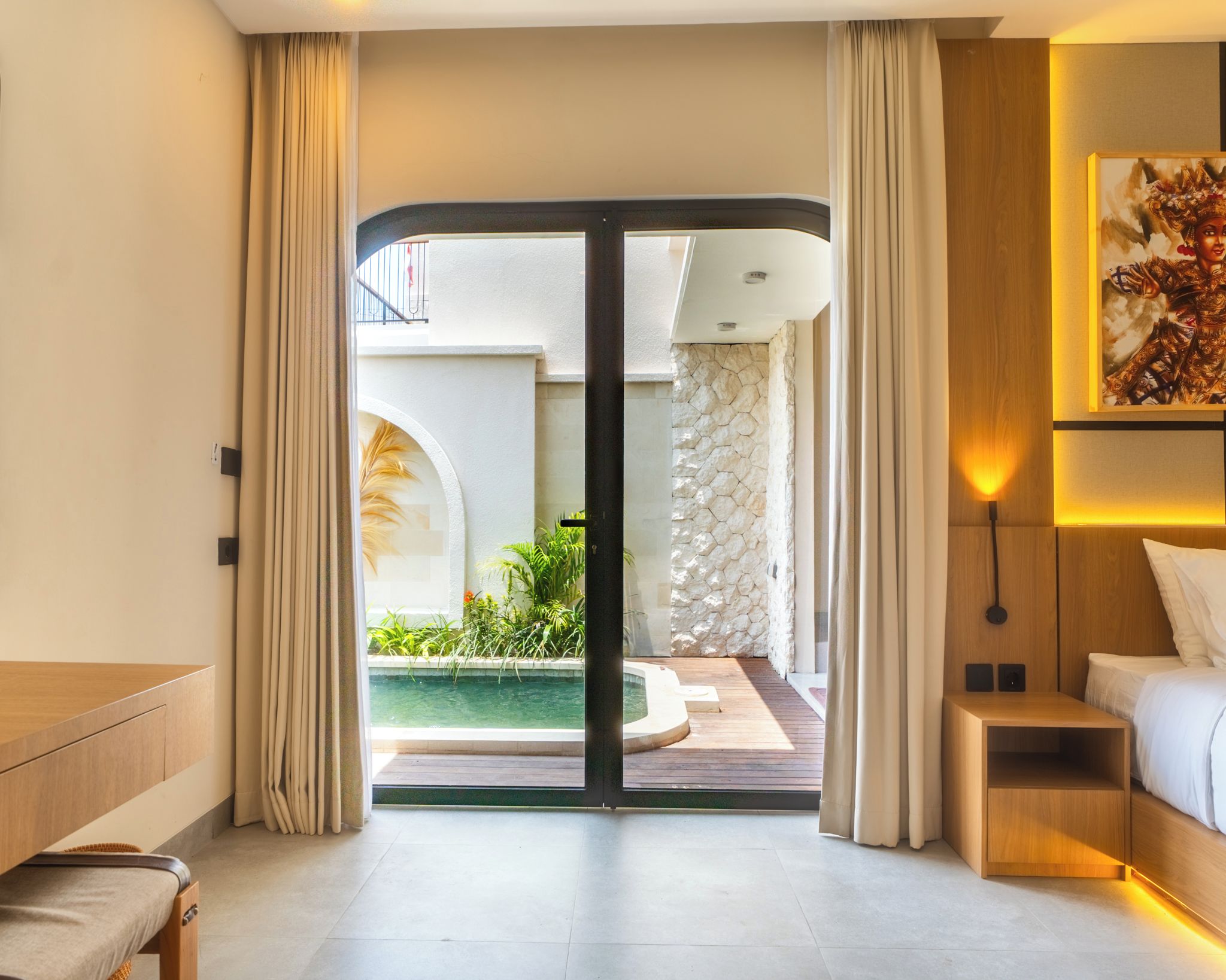 Habitación de hotel con puerta corrediza de vidrio que da a una zona de piscina, una cama, una mesita de noche y un aplique en la pared.