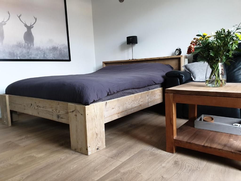 Un lit simple avec un cadre en bois et un matelas de couleur foncée, une table de chevet ornée d’un vase de fleurs et une horloge murale.