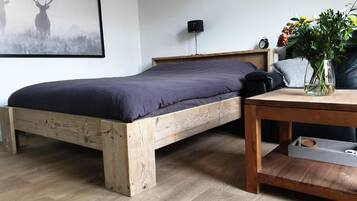 Un lit simple avec un cadre en bois et un matelas de couleur foncée, une table de chevet ornée d’un vase de fleurs et une horloge murale.