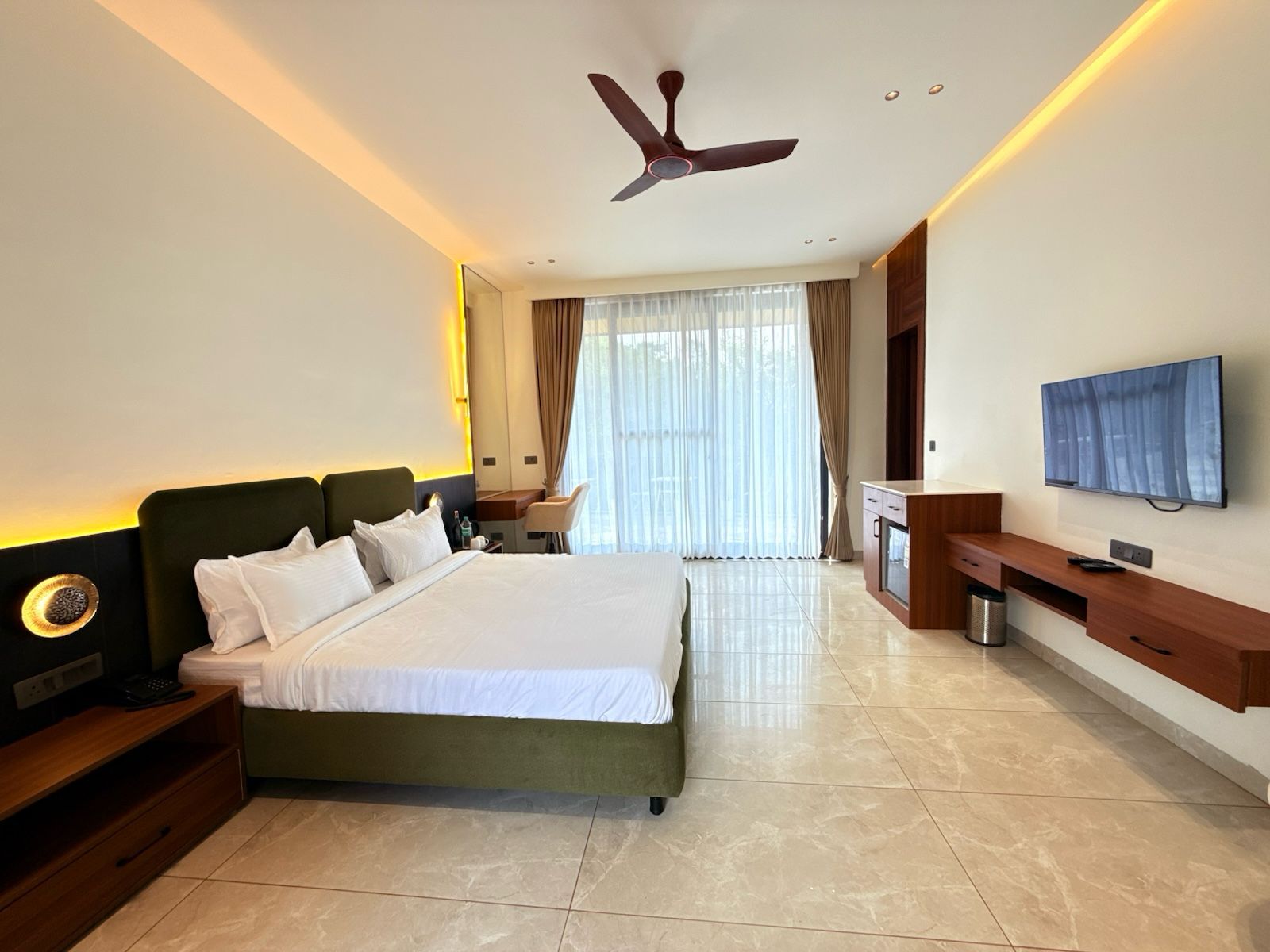 Habitación de hotel con cama, escritorio, televisor, ventilador de techo y ventana grande.
