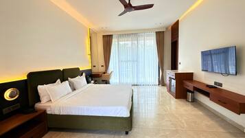 Habitación de hotel con cama, escritorio, televisor, ventilador de techo y ventana grande.