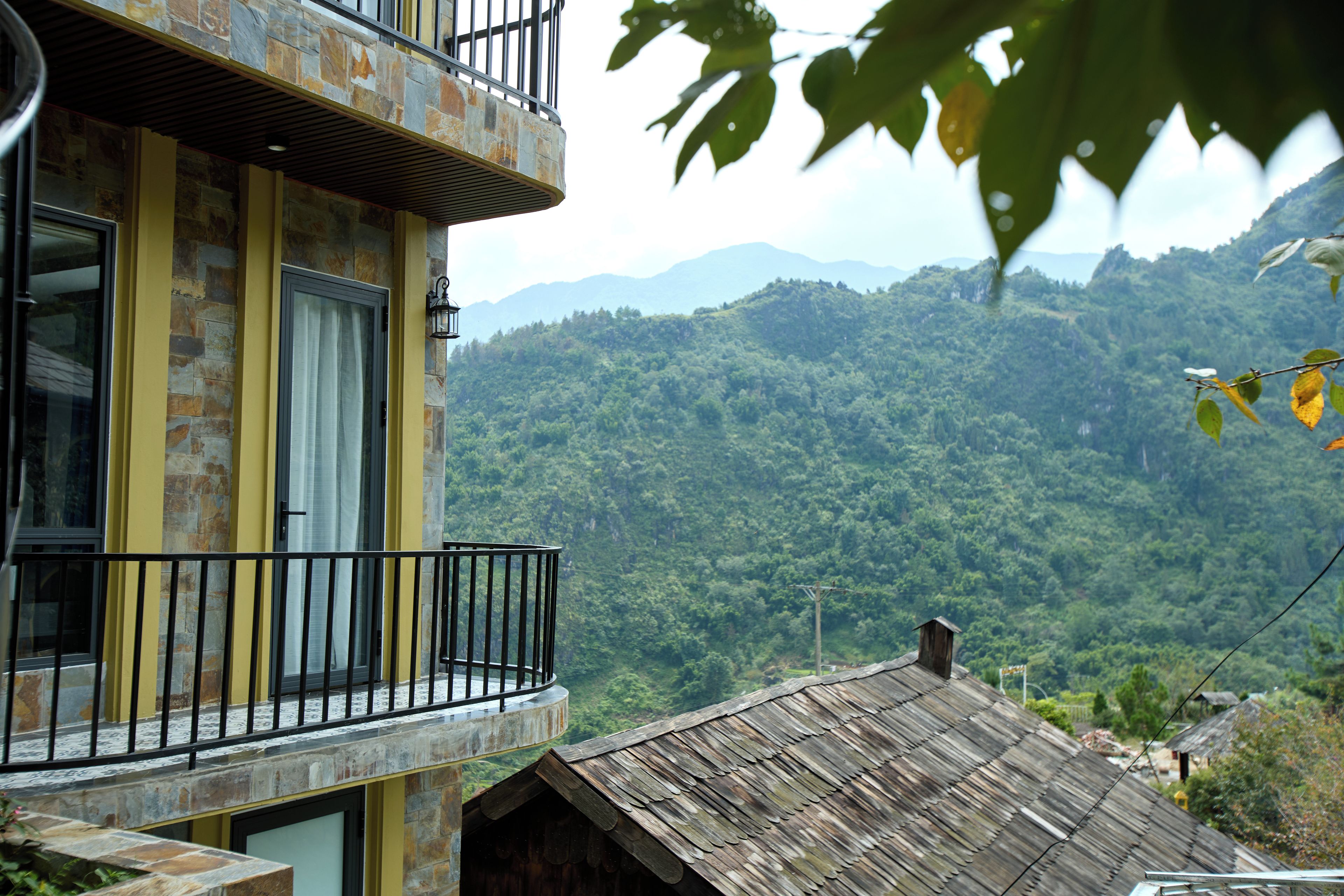 A multi-story building with a balcony, stone facade, and a view of a forested mountain range.