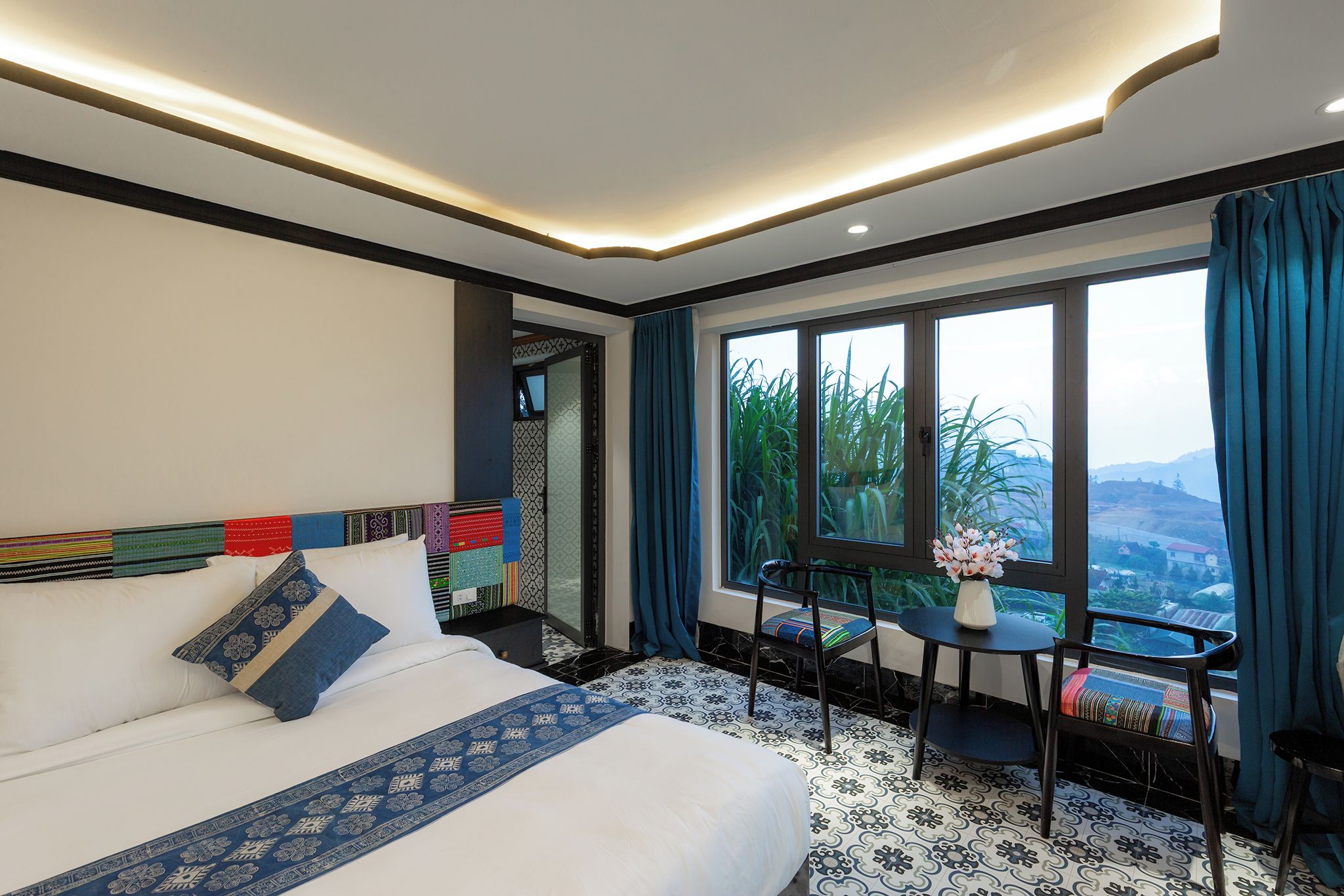 A hotel room with a bed, a small table with a vase, a chair, and a view of greenery through large windows.