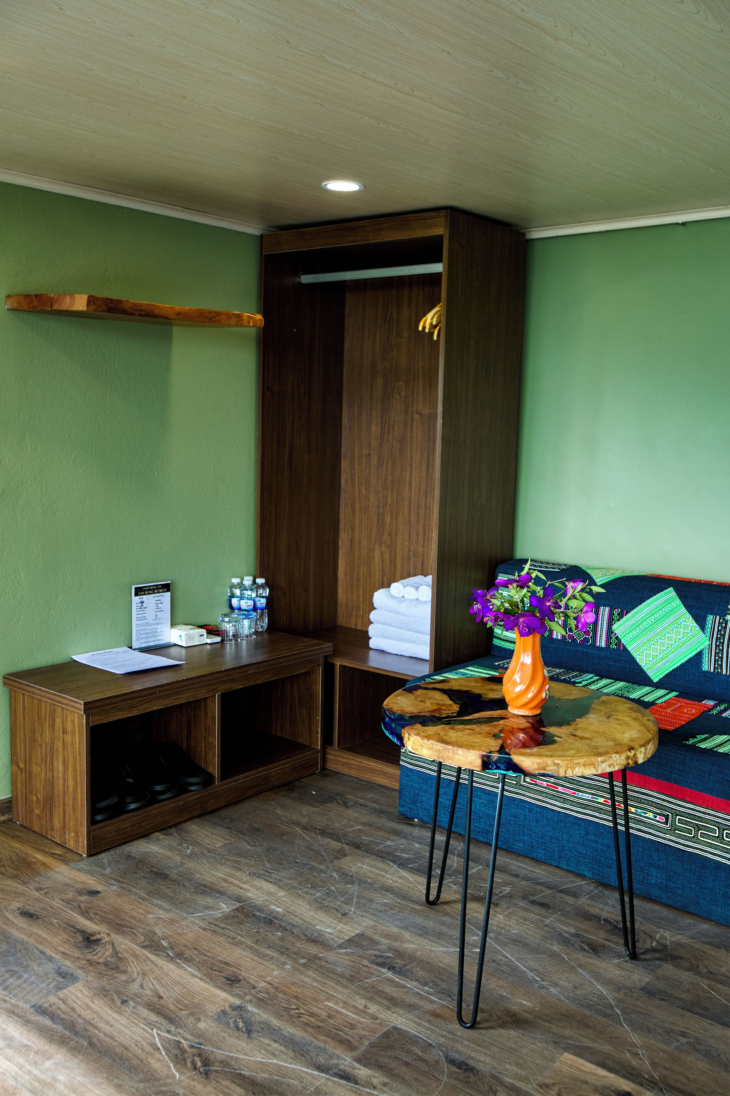 Una habitación con un escritorio de madera, una pequeña mesa redonda con un florero, un sofá con cojines estampados y un armario de madera.