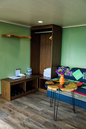 Una habitación con un escritorio de madera, una pequeña mesa redonda con un florero, un sofá con cojines estampados y un armario de madera.