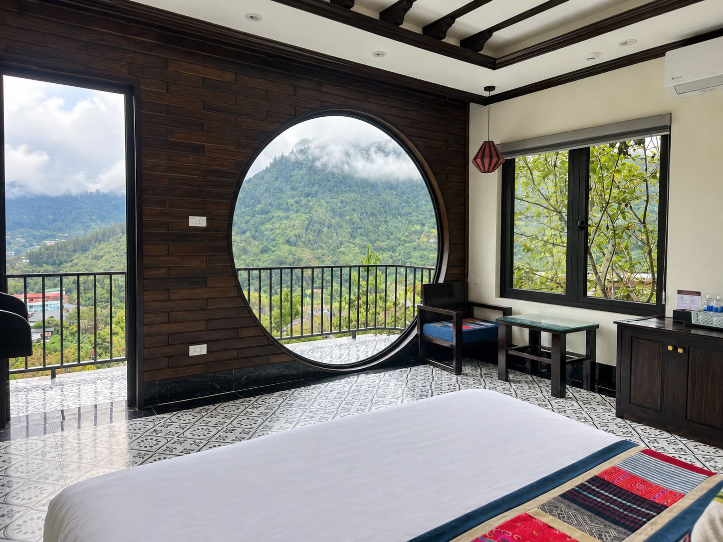 A bedroom with a large bed, a circular window, a balcony, and a view of a mountainous landscape.