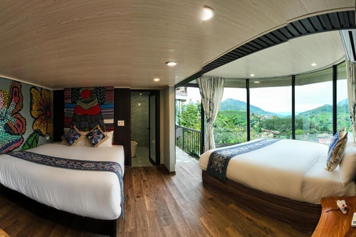 A hotel room with two beds, a mural on the wall, and a balcony with a view of the mountains.
