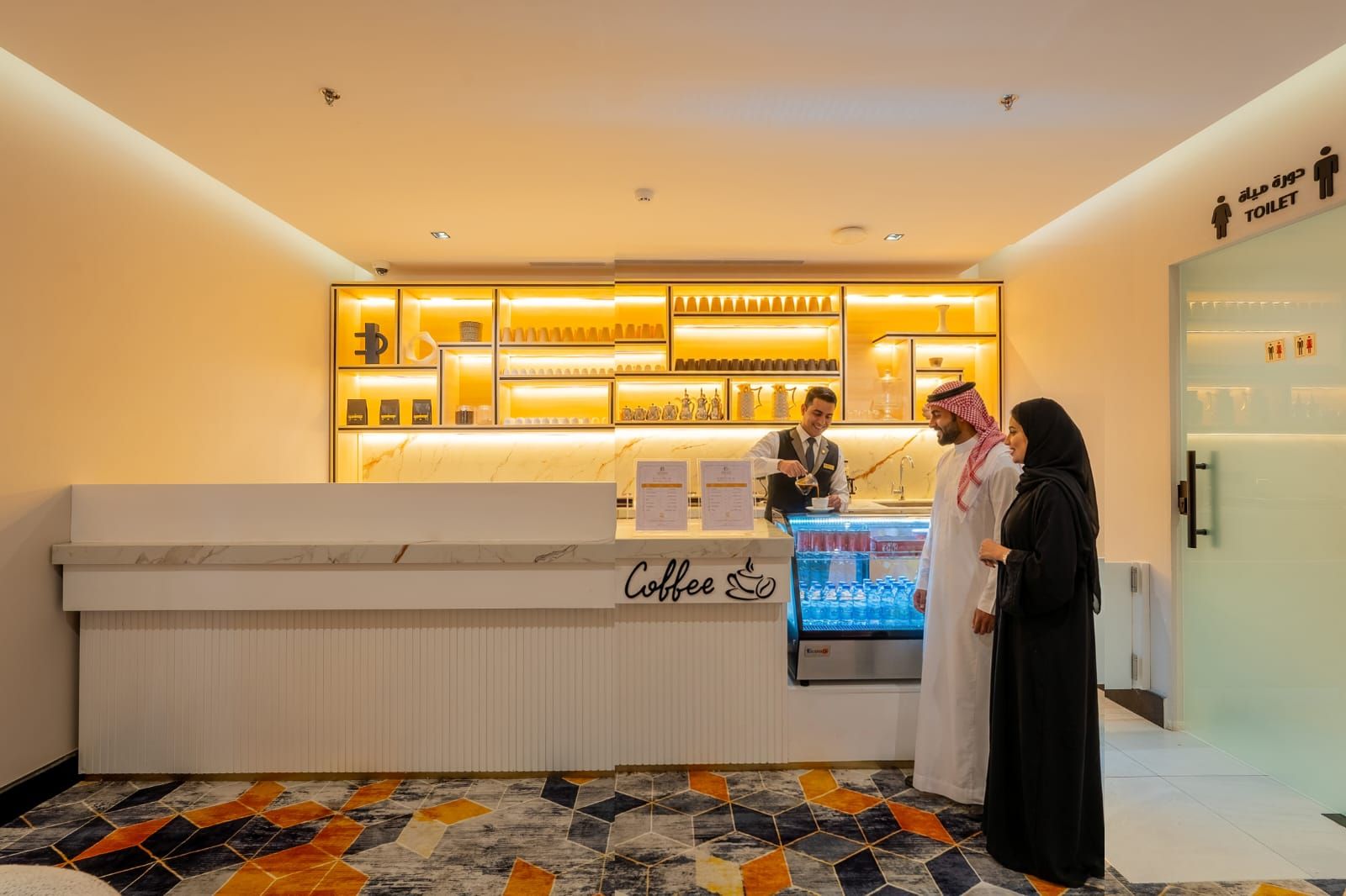 A coffee bar with a marble countertop and a refrigerated display, a sign indicating a toilet, and a patterned floor.