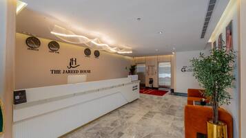 A hotel reception area with a reception desk, clocks displaying different cities, and a red carpet leading to elevators.