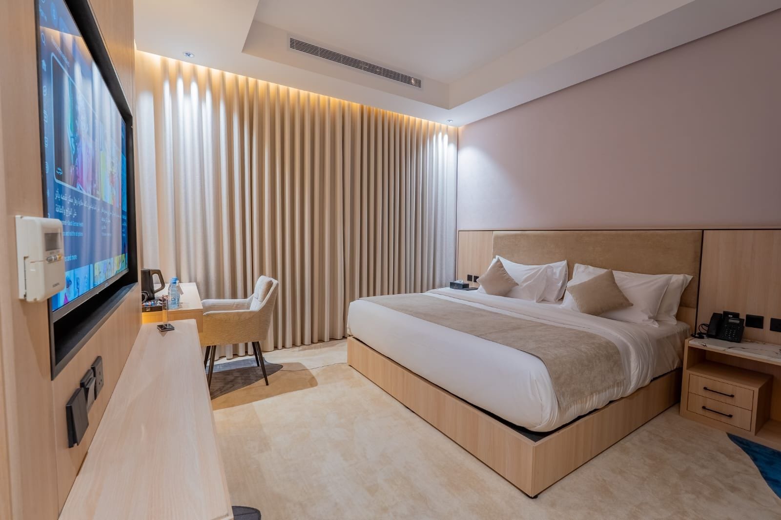 Habitació d'hotel moderna amb un llit gran, una taula de treball amb una cadira, una televisió i un panell de control fixat a la paret.