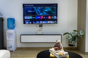 Ein modernes Wohnzimmer mit einem an der Wand befestigten Fernseher, der eine Streaming-Oberfläche zeigt, einem Wasserspender, einer Zimmerpflanze und einem einzigartigen Mittelstück auf einem Tisch.