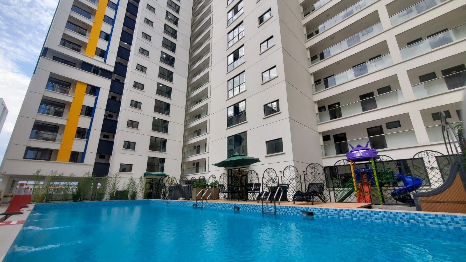 A swimming pool with a slide and lounge chairs, surrounded by a tall residential building with balconies.