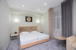 Een hotelkamer met een bed, nachtkastjes, een kleintje tafeltje, een stoel en een raam met gordijnen.