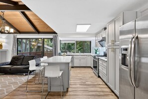 Een moderne keuken met roestvrijstalen apparatuur, een eethoek met een houten tafel en stoelen, en uitzicht op het omliggende landschap door grote ramen.