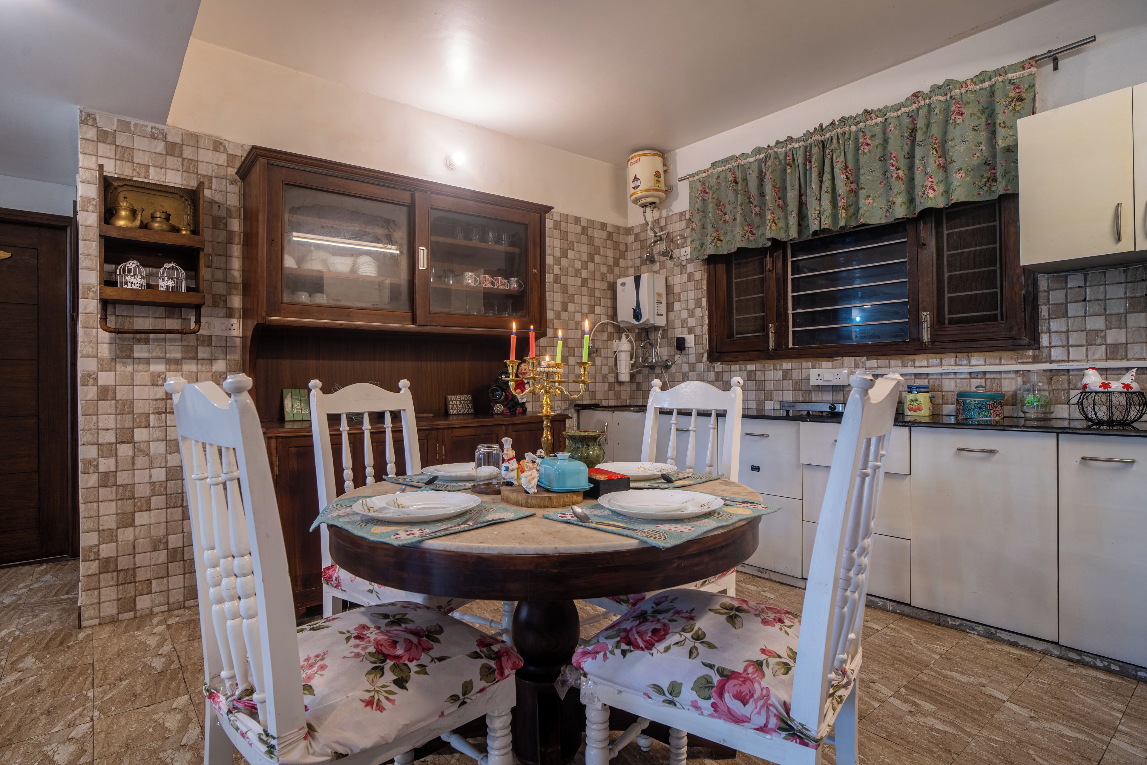 Comedor con mesa redonda, sillas blancas con tapicería floral y una cocina al fondo con armarios de madera y salpicadero de azulejos.