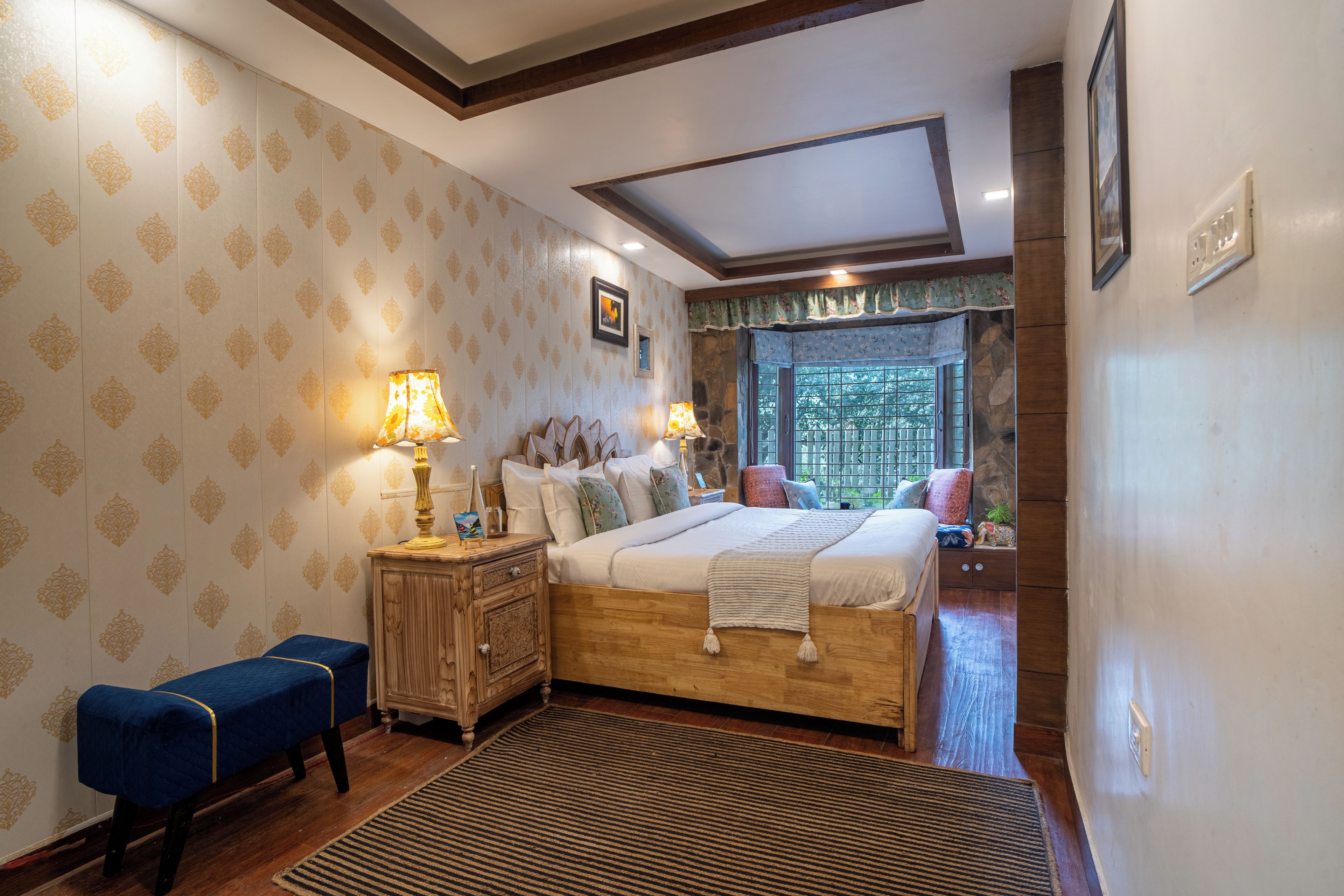 A bedroom with a large bed, a nightstand, a bench, and a window with a view of trees.