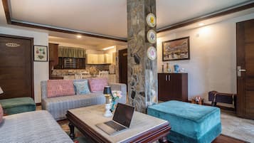 A living room with a sofa, a coffee table with a laptop, a stone pillar, and a kitchen in the background.