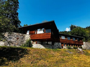 Une maison moderne à deux étages, dotée d’une façade en pierre et de détails en bois, située dans un paysage verdoyant et luxuriant.