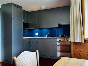 Eine kleine Küche mit grauen Schränken, einer blauen Spritzschutzwand, einem Spülbecken, einem Herd und einem Holztisch.