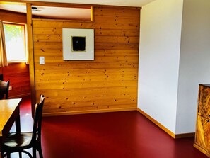 Ein Zimmer mit Holzwandverkleidung, rotem Holzboden, einer weißen Wand mit einem gerahmten Bild, einem Holzschrank und einem Esstisch mit Stühlen.