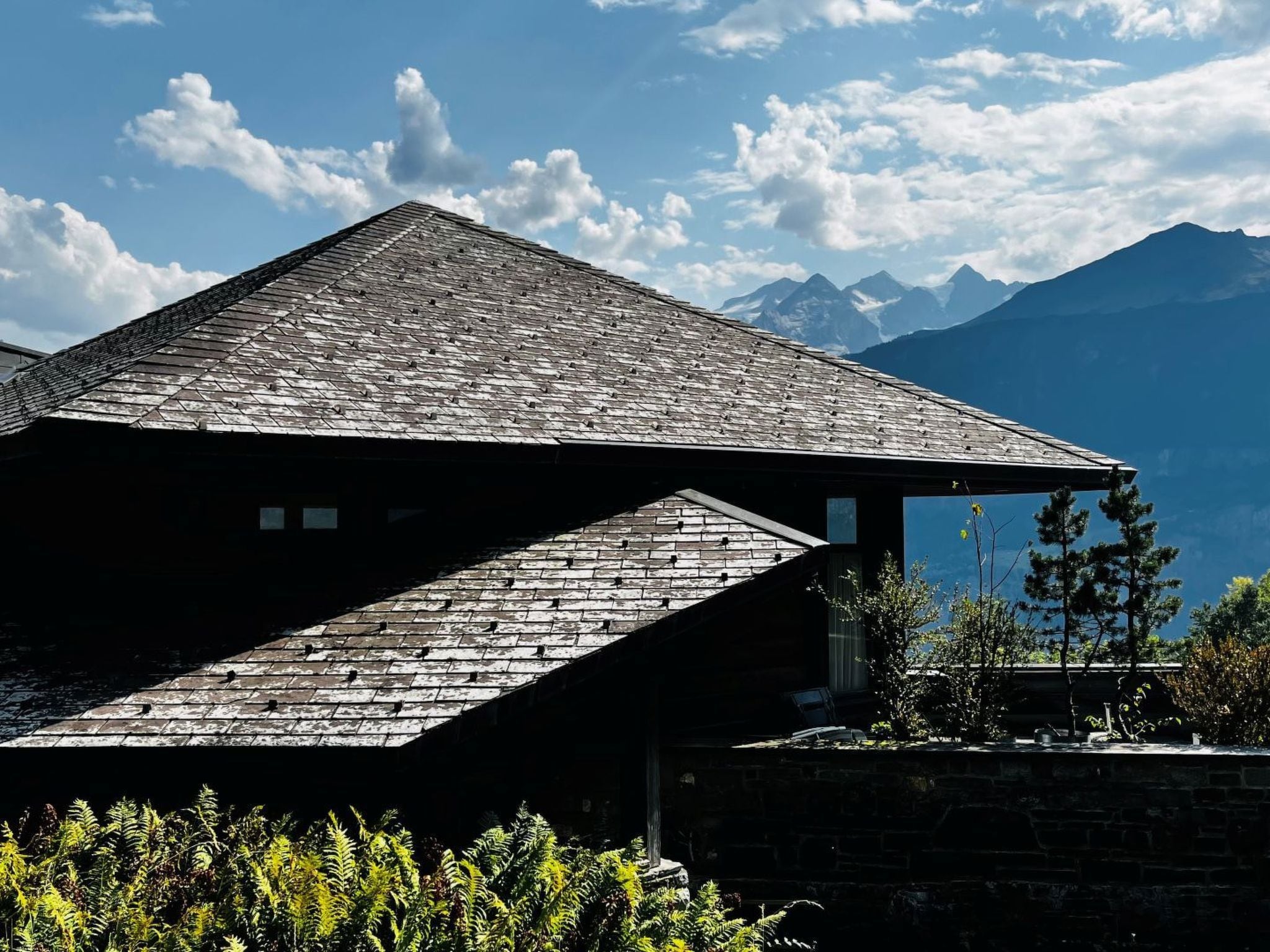 A building with a sloped roof, a stone wall, and a view of mountains in the background.