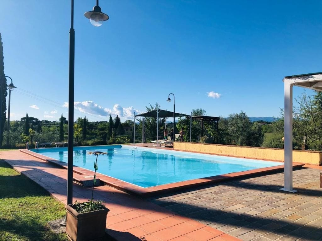 Una piscina rectangular con borde de azulejos, rodeada por un patio de ladrillo. Se observa una estructura de pérgola con techo al fondo.