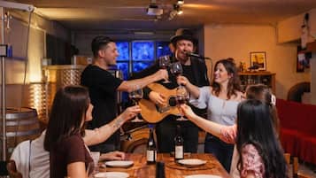 Un groupe de personnes trinquant avec des verres de vin autour d’une table en bois, dans une pièce où se trouvent un canapé rouge et une guitare.