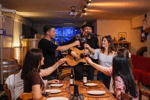Um grupo de pessoas brindando com taças de vinho ao redor de uma mesa de madeira em um quarto com sofá vermelho e um violão.