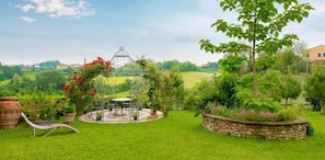 A garden with a gazebo, a table, and chairs, surrounded by various plants and a stone wall.