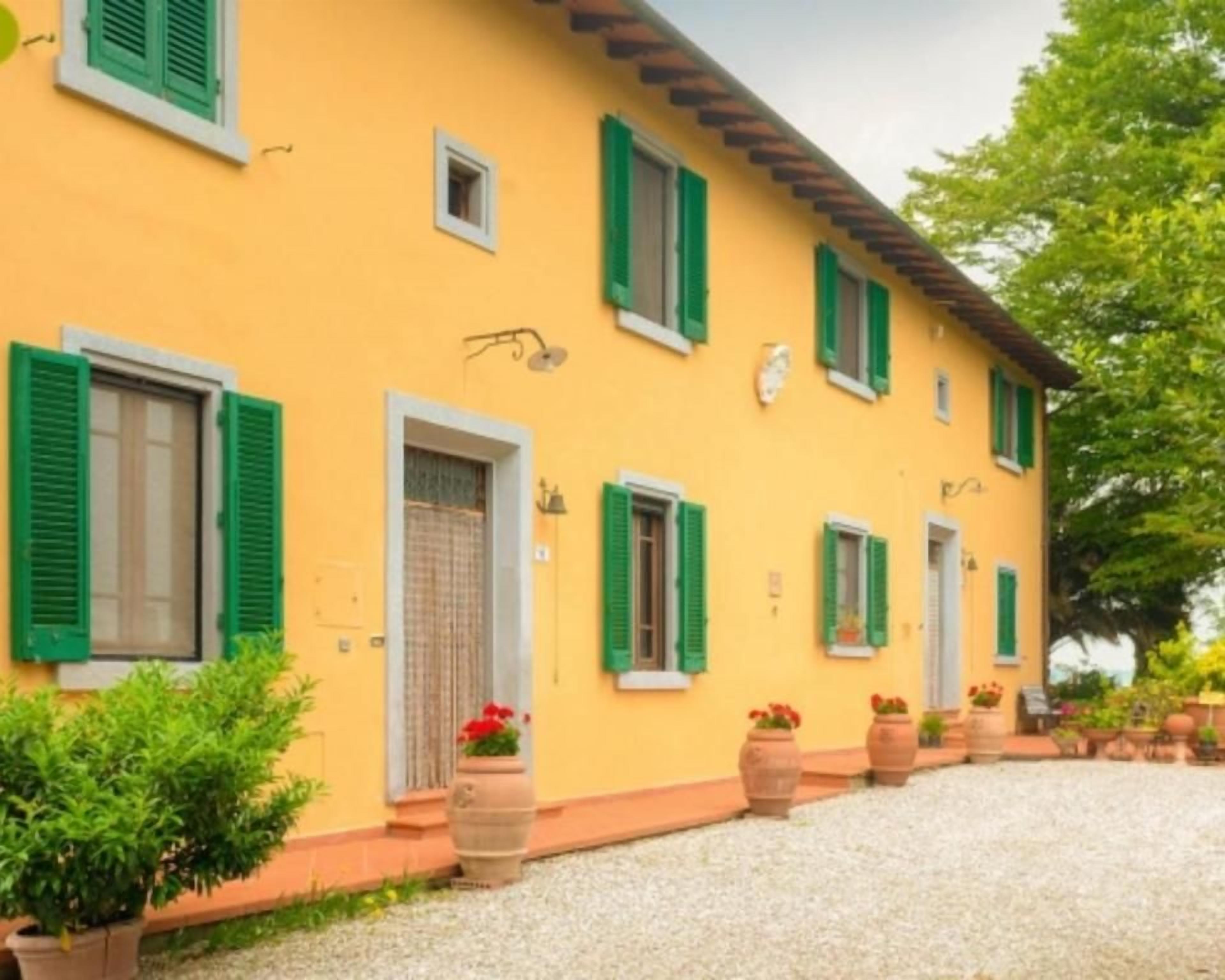 Um edifício amarelo com persianas verdes, entrada com calçada de cascalho e vasos de plantas.